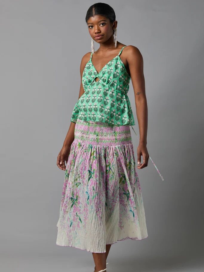 A woman stands against a gray background wearing a green floral sleeveless top, a pleated midi skirt with pink and purple patterns, and white strappy sandals.