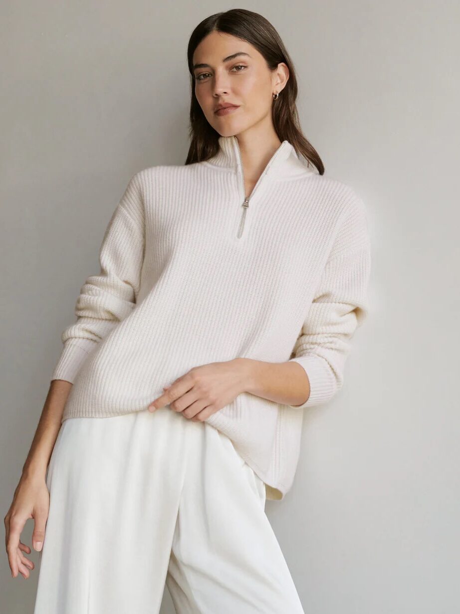 A woman stands against a plain background wearing a cream-colored zip-neck sweater and loose white pants, with one hand resting on her hip.