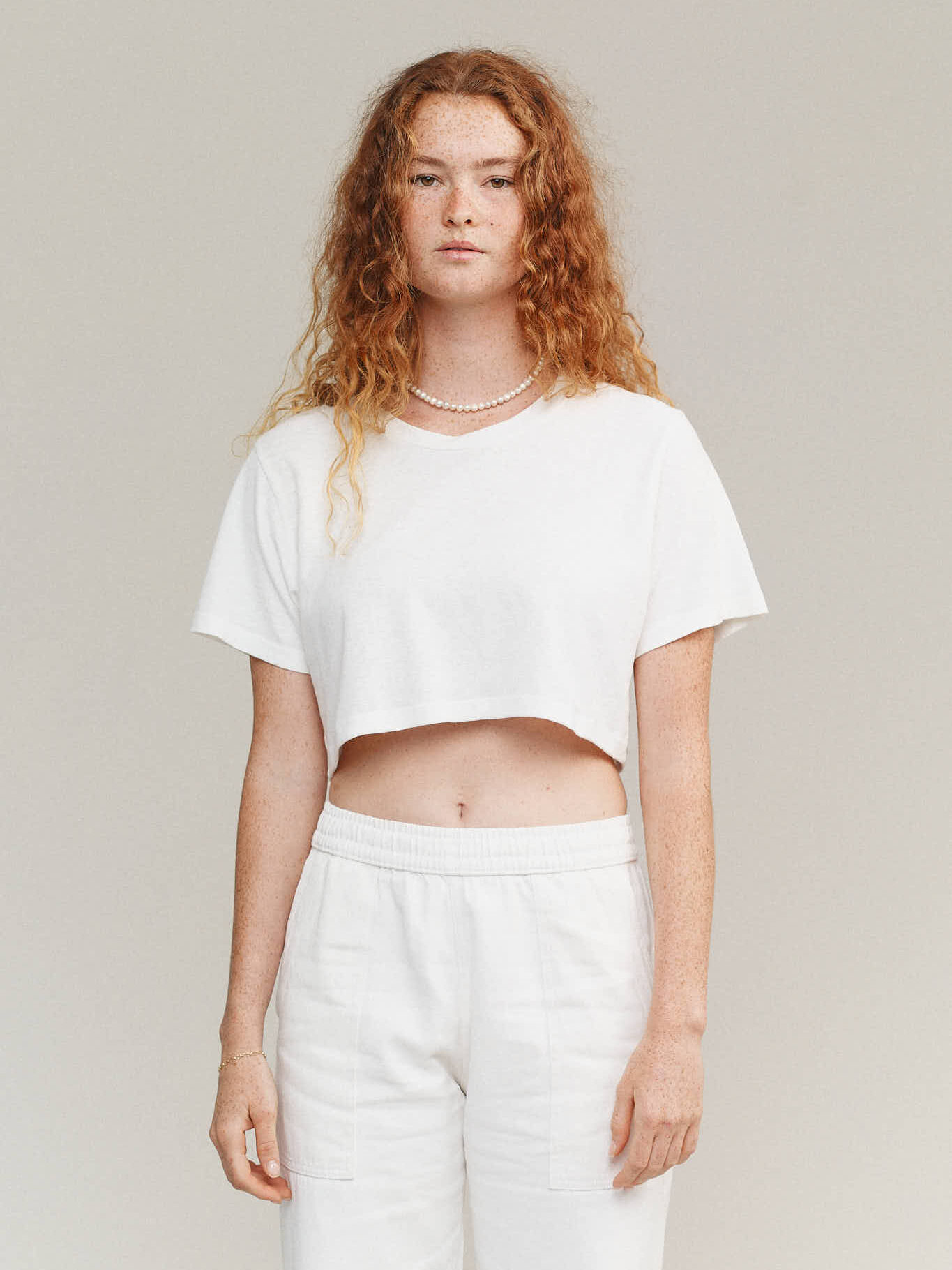 A person with long curly hair wears a white cropped t-shirt, white pants, and a pearl necklace, standing against a plain light background.