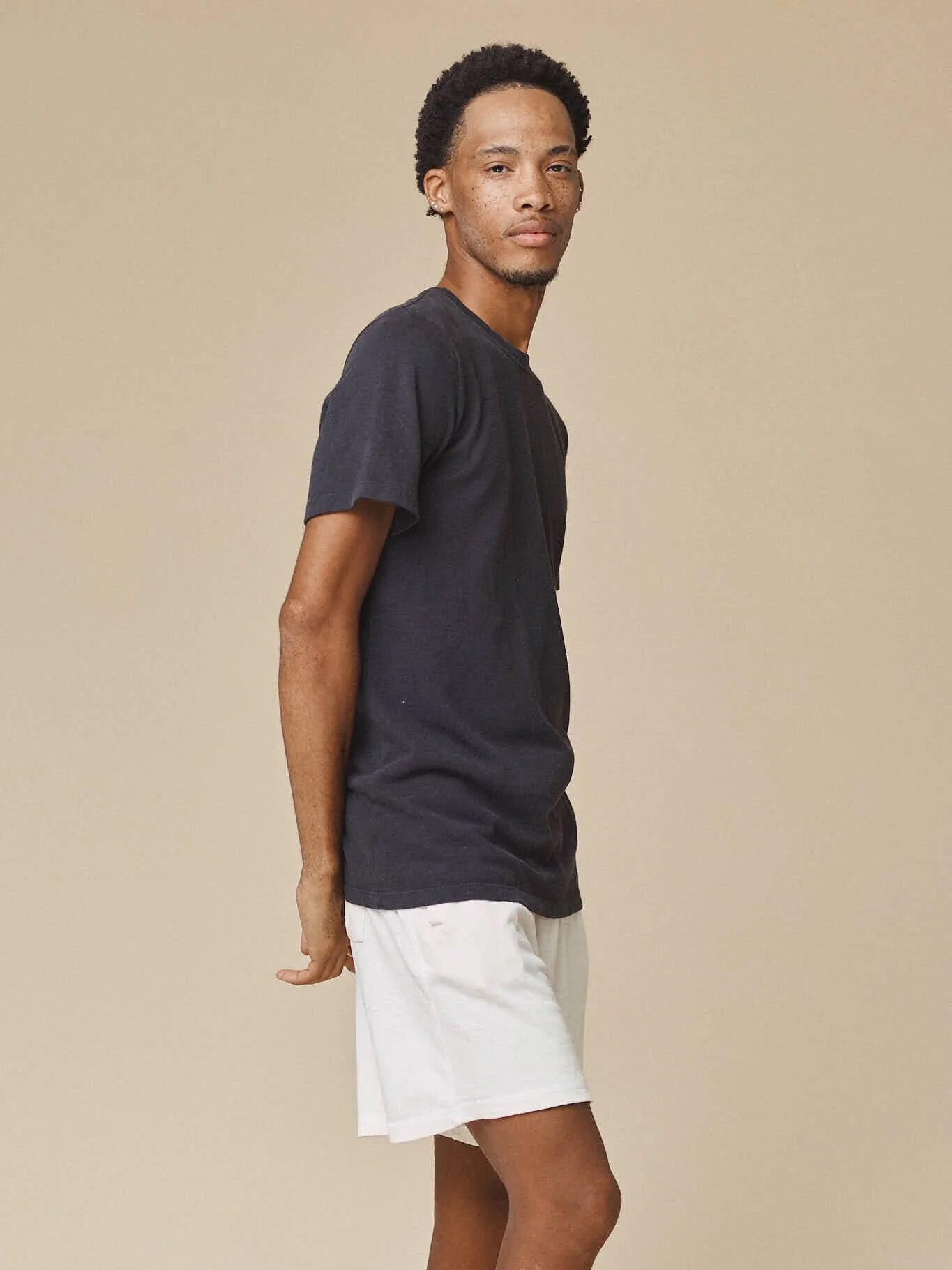 A man stands sideways against a plain beige background, wearing a black t-shirt and white shorts.