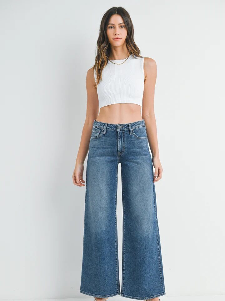 Woman standing against a white background wearing a white sleeveless crop top, high-waisted wide-leg blue jeans, and open-toe sandals.
