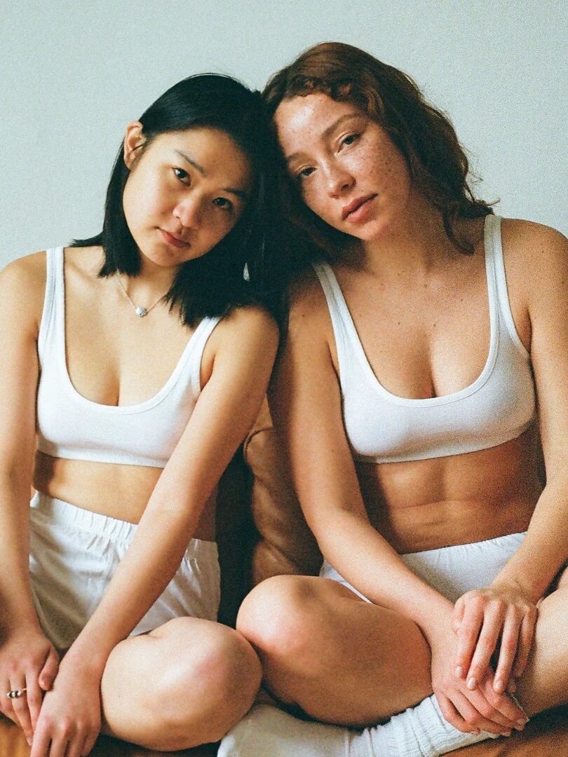 Two women sit close together on a couch, both wearing white sports bras and shorts, looking at the camera with relaxed expressions.