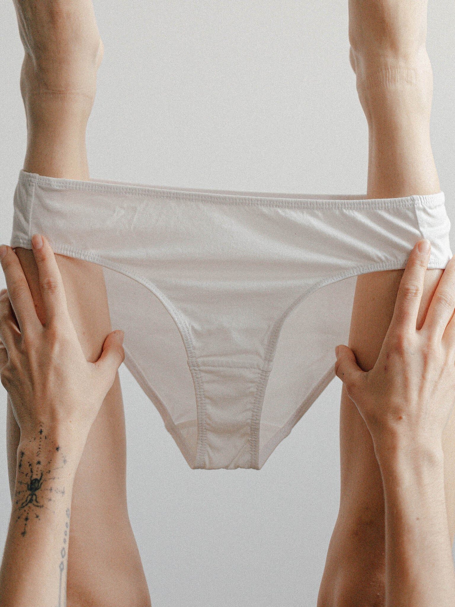 A person holds up their legs vertically while pulling on a pair of white underwear, with hands visible and a tattoo on one wrist.