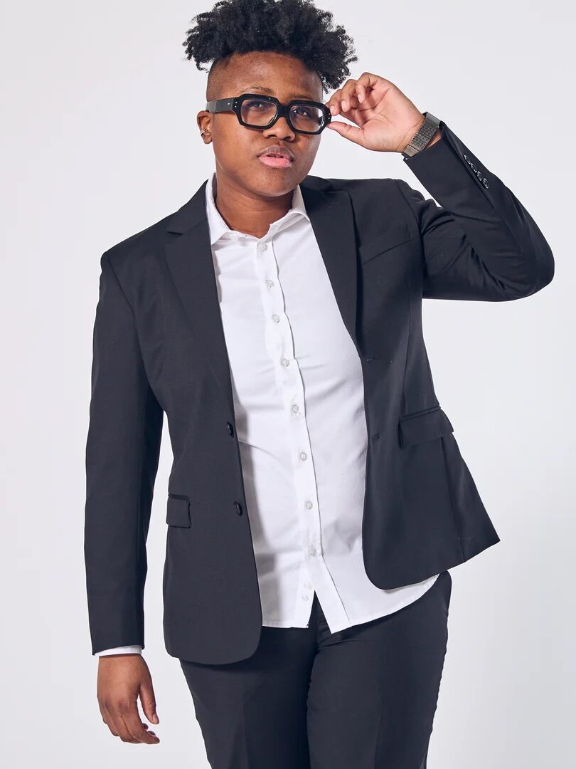 Person wearing a black suit and white shirt stands against a plain background, adjusting black-rimmed glasses with one hand.