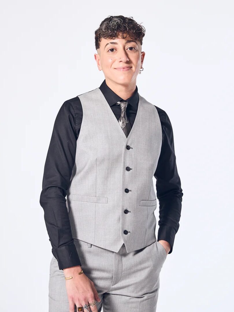 Person wearing a light gray vest and pants with a black shirt and patterned tie, standing against a plain white background with one hand in their pocket.