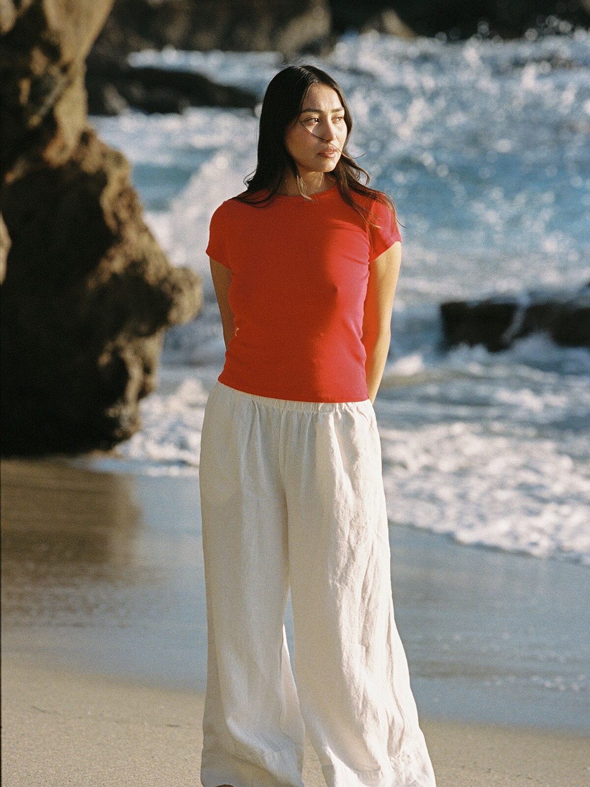 A woman in a red shirt and white pants stands on a sandy beach near rocks, with waves and sunlight in the background.
