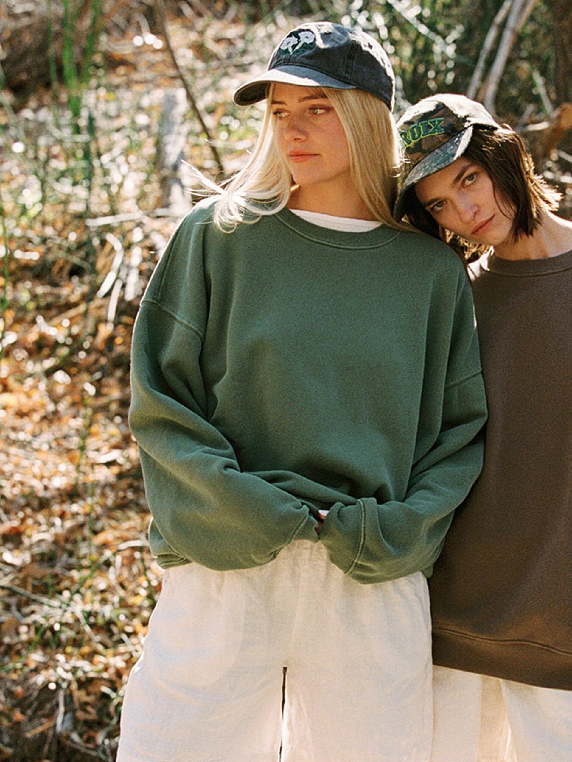 Two people outdoors wear oversized sweatshirts, light shorts, and baseball caps. One person leans their head on the other's shoulder. Dry leaves and trees are visible in the background.