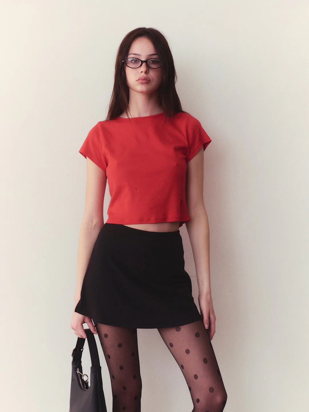 A woman wearing glasses, a red crop top, a black skirt, and polka dot tights stands against a light-colored wall, holding a black handbag.