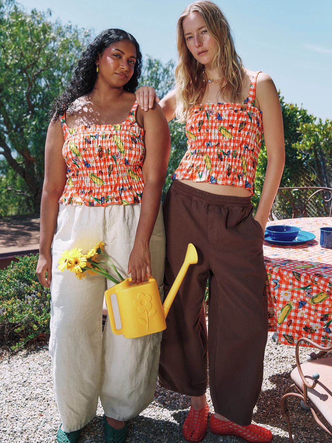 Two women stand outdoors wearing patterned orange tops; one holds a yellow watering can and sunflowers, both are dressed in loose pants. A table with a checkered tablecloth is in the background.