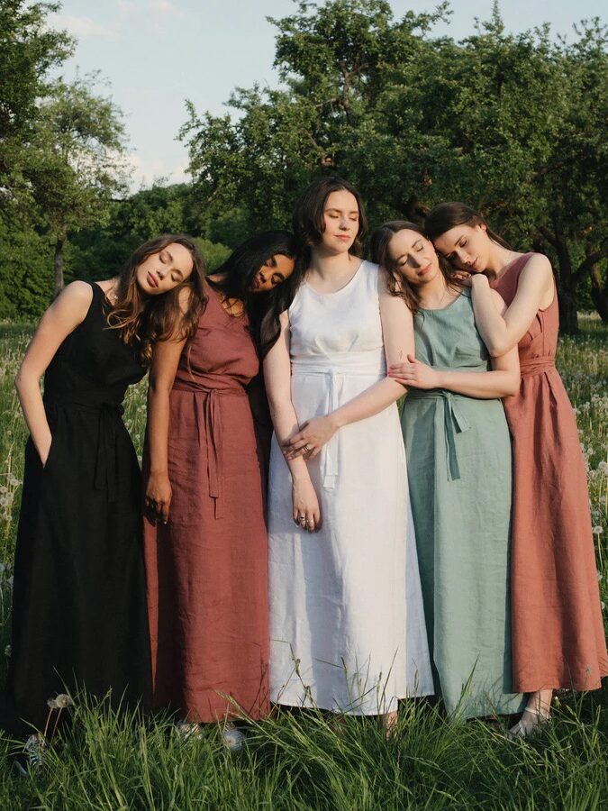 Five women in sleeveless, belted dresses stand closely together outdoors in a grassy area with trees, some leaning on each other and resting heads on shoulders.