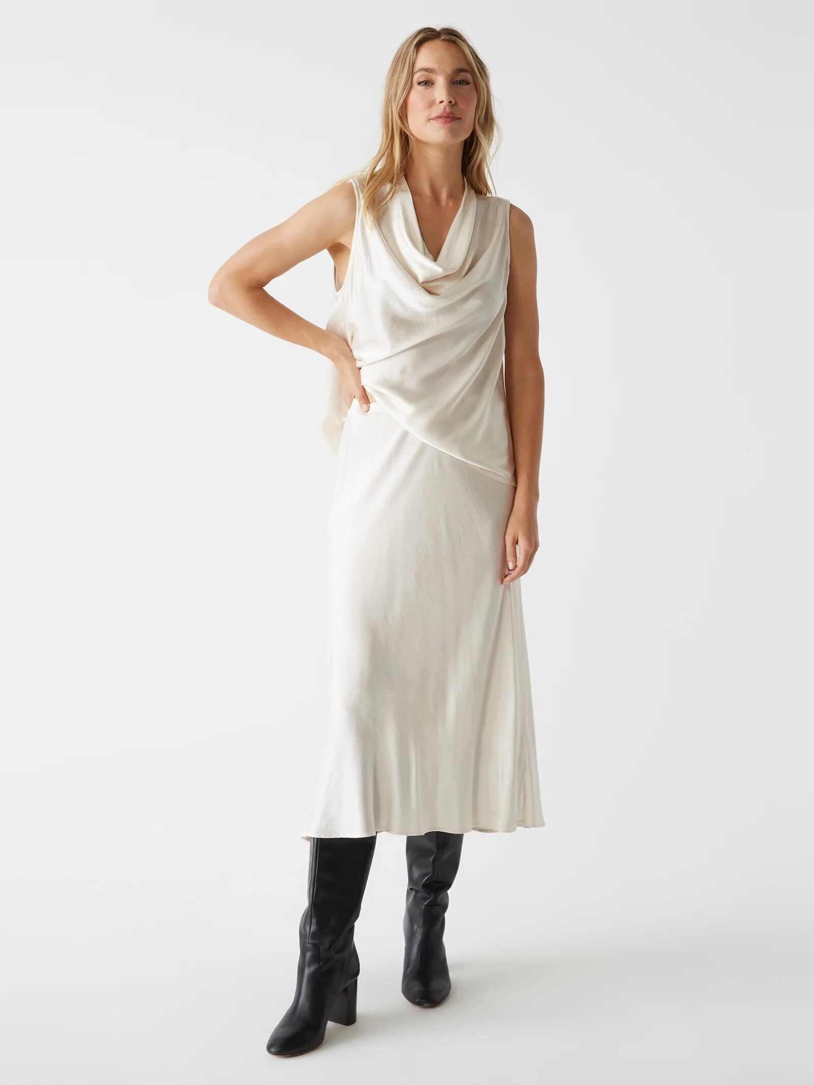 A woman stands against a plain background wearing a sleeveless, ivory cowl-neck midi dress and black knee-high boots.