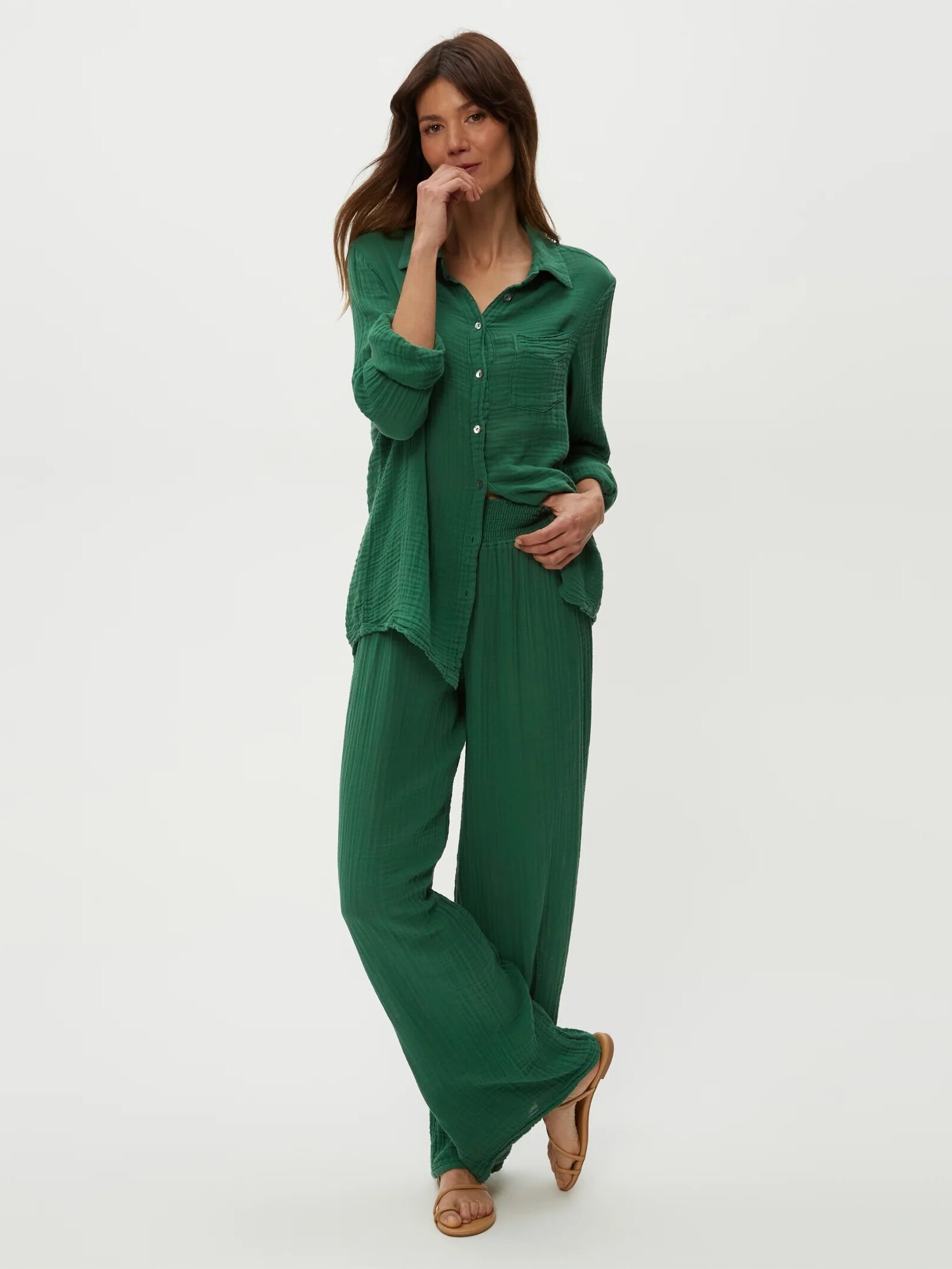 Woman standing against a plain light background, wearing a loose-fitting green button-up shirt and matching green wide-leg pants with tan sandals.