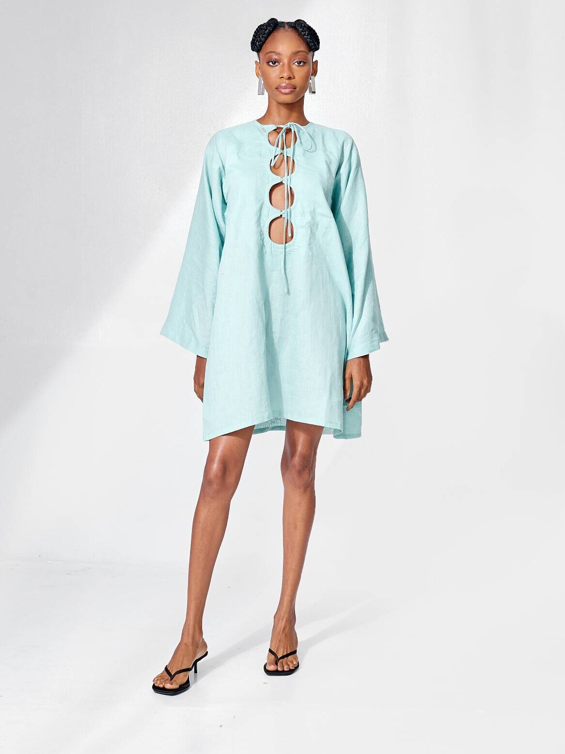 A woman stands wearing a light blue, long-sleeve dress with front cutouts and tie detail, paired with black heeled sandals, against a plain white background.
