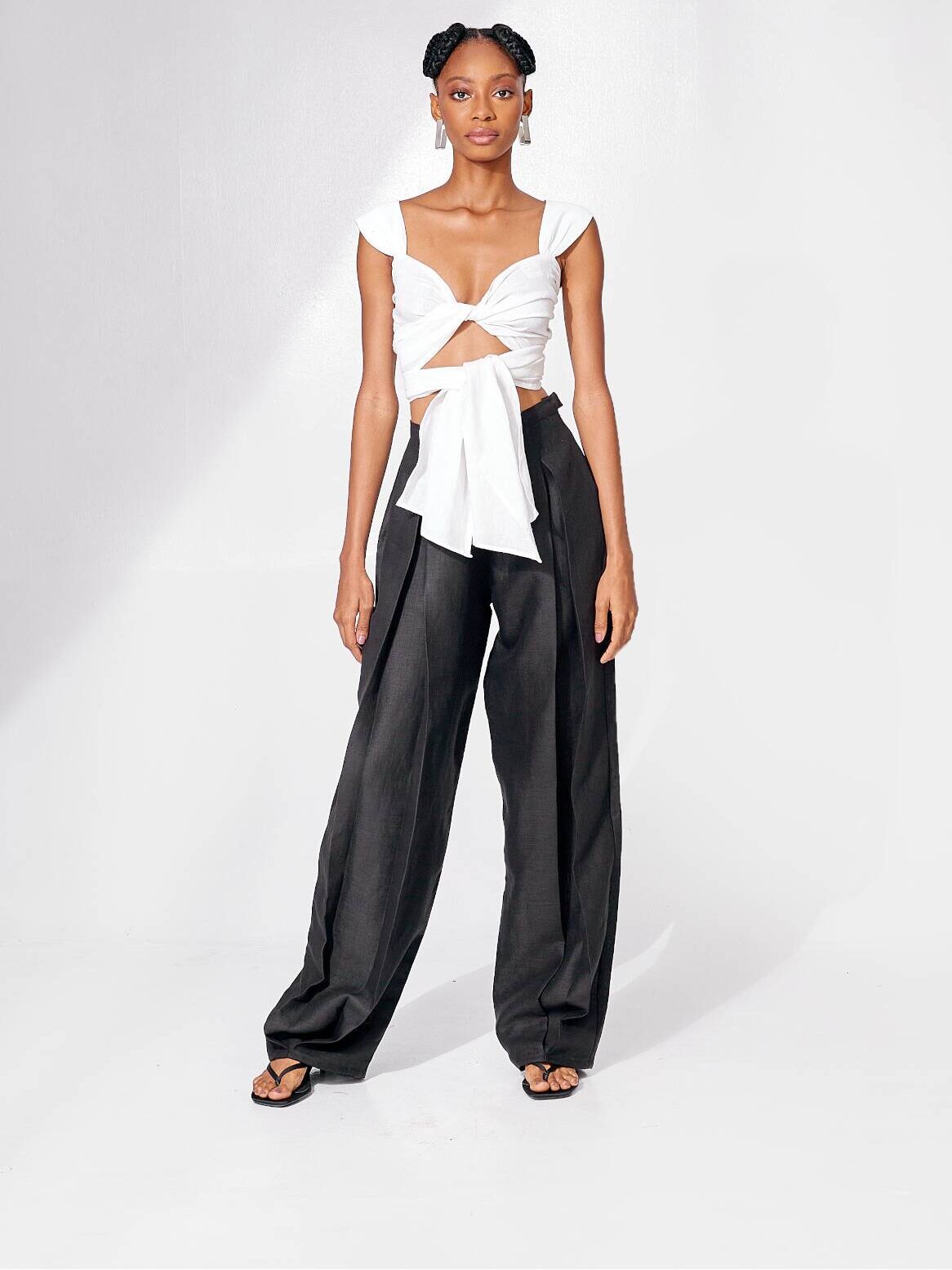 A person stands against a plain white background wearing a white knotted crop top, black wide-leg trousers, and black sandals.