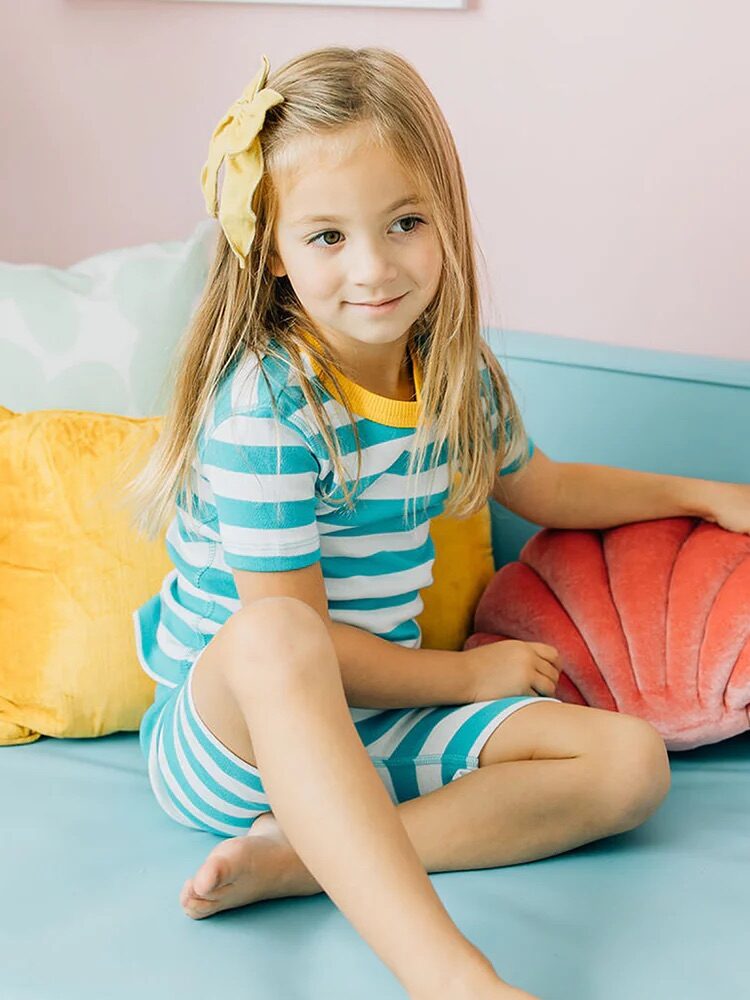 A young girl with a yellow bow sits on a light blue couch, wearing blue and white striped pajamas, with yellow and pink pillows beside her.