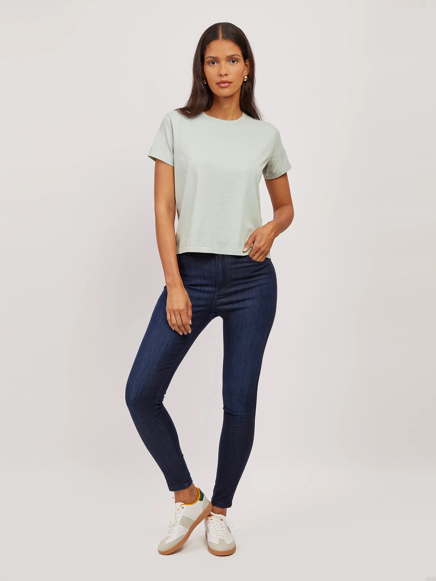 A woman stands facing forward, wearing a light green short-sleeve t-shirt, dark blue skinny jeans, and white sneakers, against a plain light background.