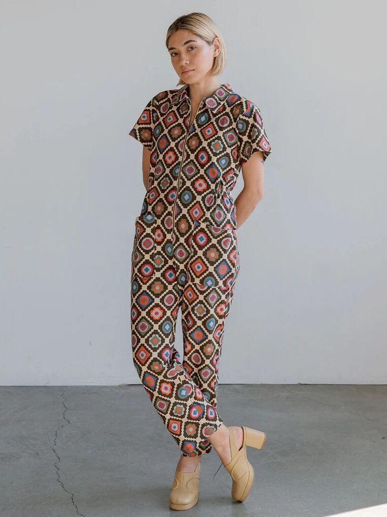 A person stands indoors against a plain wall, wearing a patterned jumpsuit with short sleeves and tan heeled shoes, with hands in their pockets.