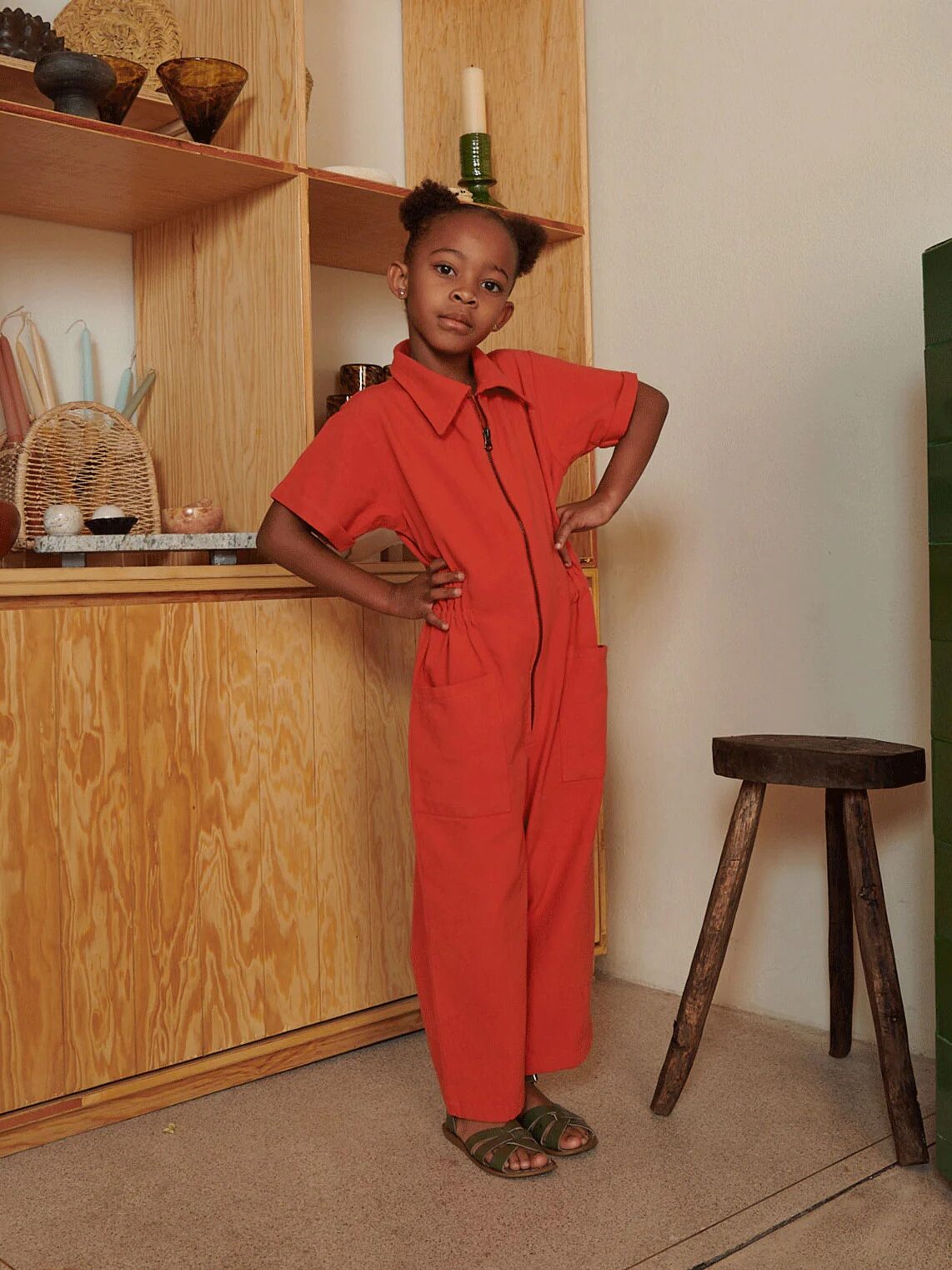 A young girl stands indoors wearing a red jumpsuit and green sandals, with one hand on her hip, next to a wooden stool and shelves displaying various objects.