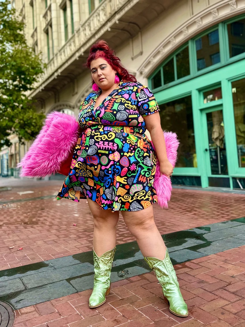 A person stands on a wet brick sidewalk wearing a colorful patterned dress, metallic green boots, and holding a pink furry accessory, with buildings in the background.