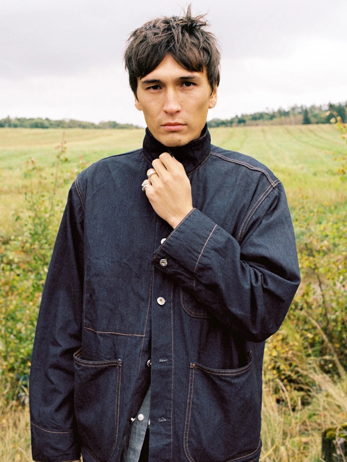 A person stands outdoors in a grassy field wearing a dark denim jacket, holding the collar, and looking directly at the camera.