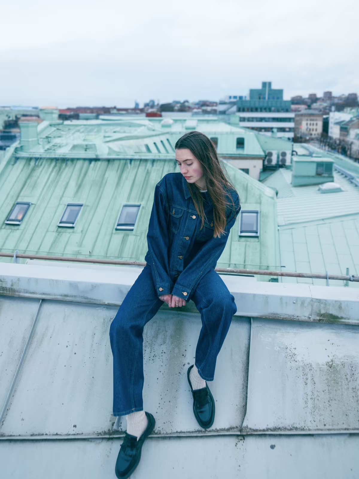 A woman in a denim jacket and jeans sits on a rooftop ledge, looking down, with green rooftops and a cityscape in the background.
