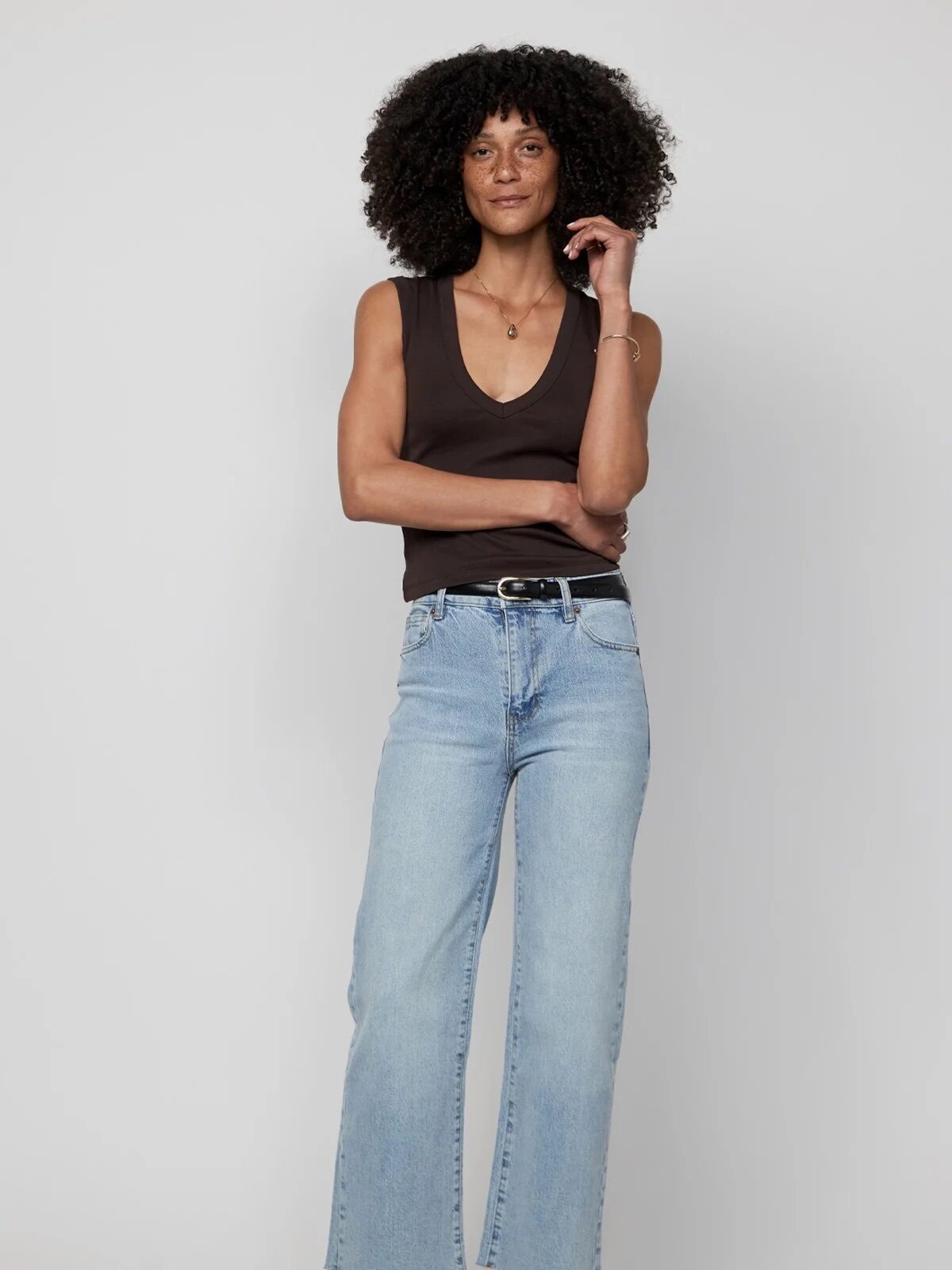 A person stands against a plain background, wearing a sleeveless black top, light blue jeans, and black shoes, with one hand touching their hair.