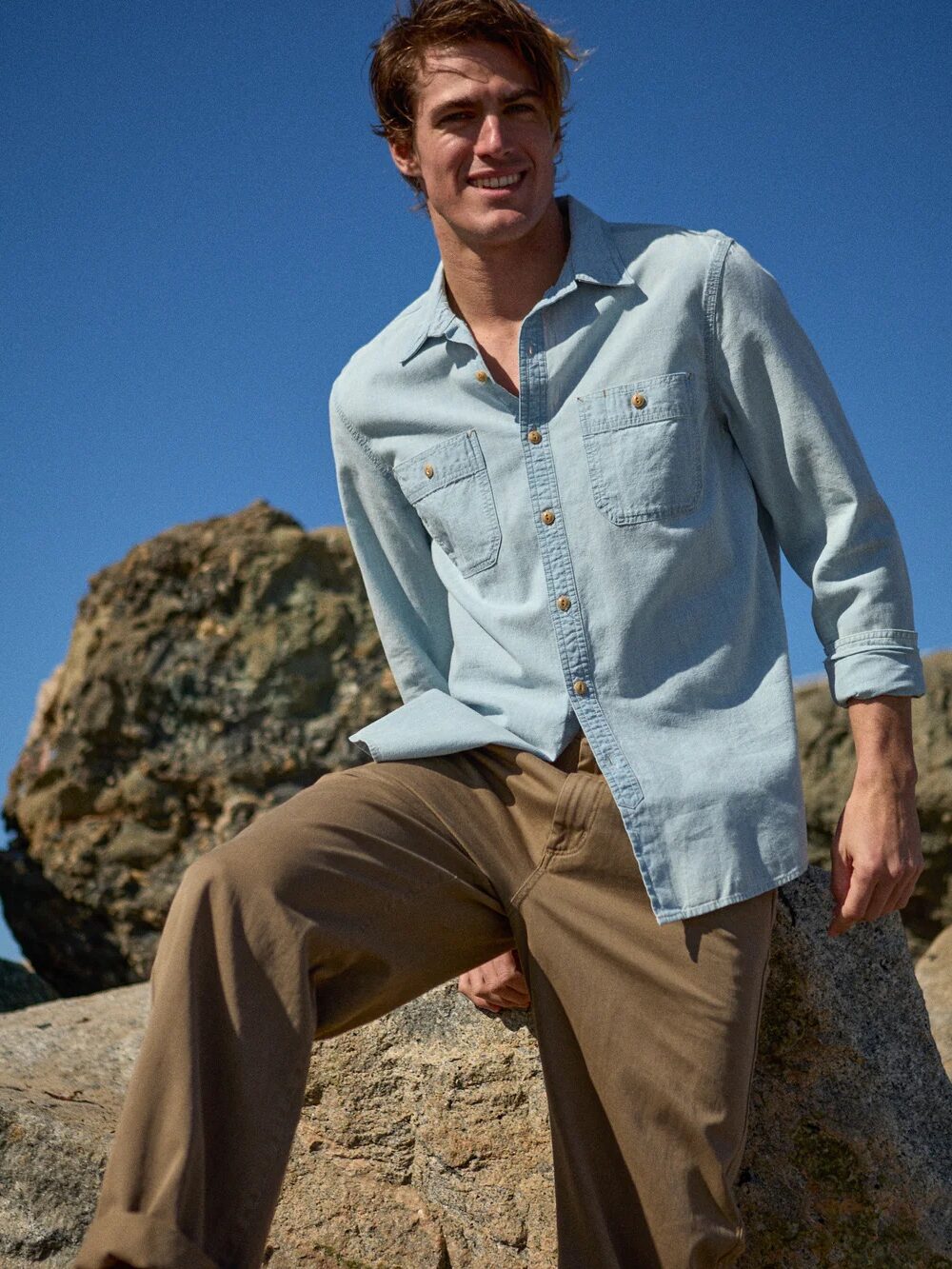 A man wearing a light denim shirt and khaki pants sits on a rock outdoors with large rocks and a clear blue sky in the background.