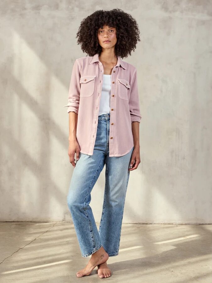 A woman with curly hair stands barefoot indoors, wearing a light pink button-up shirt over a white top and blue jeans, against a plain, light-colored wall.