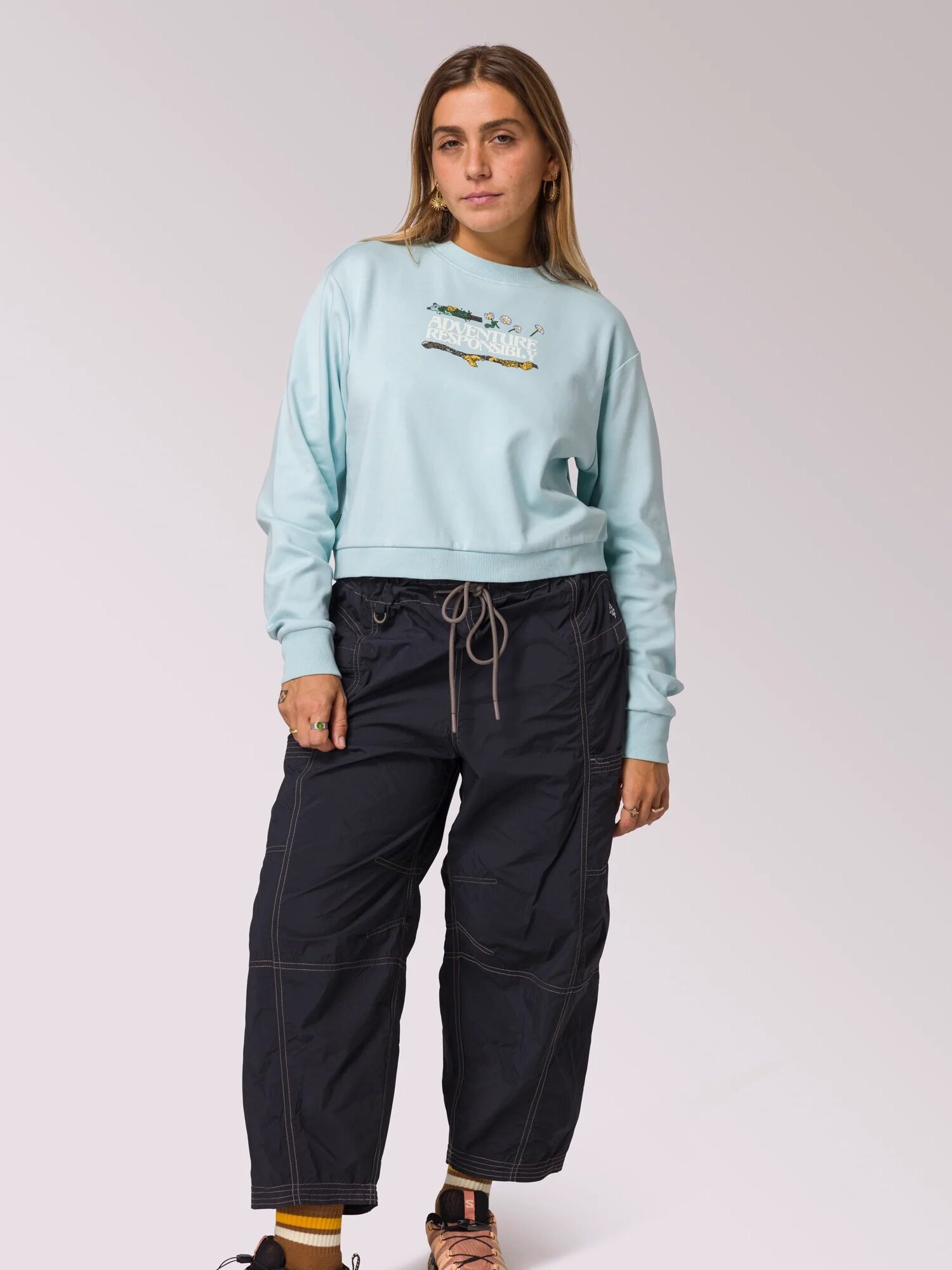 A woman stands facing forward wearing a light blue sweatshirt, black drawstring pants, yellow socks, and black sandals against a plain background.