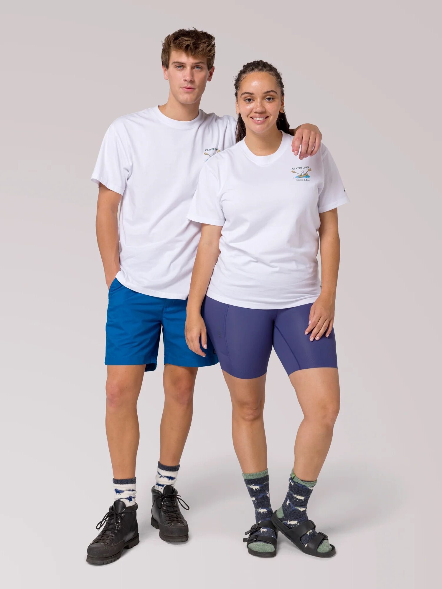 Two people stand side by side wearing white t-shirts, blue shorts, patterned socks, and sandals against a plain light background.