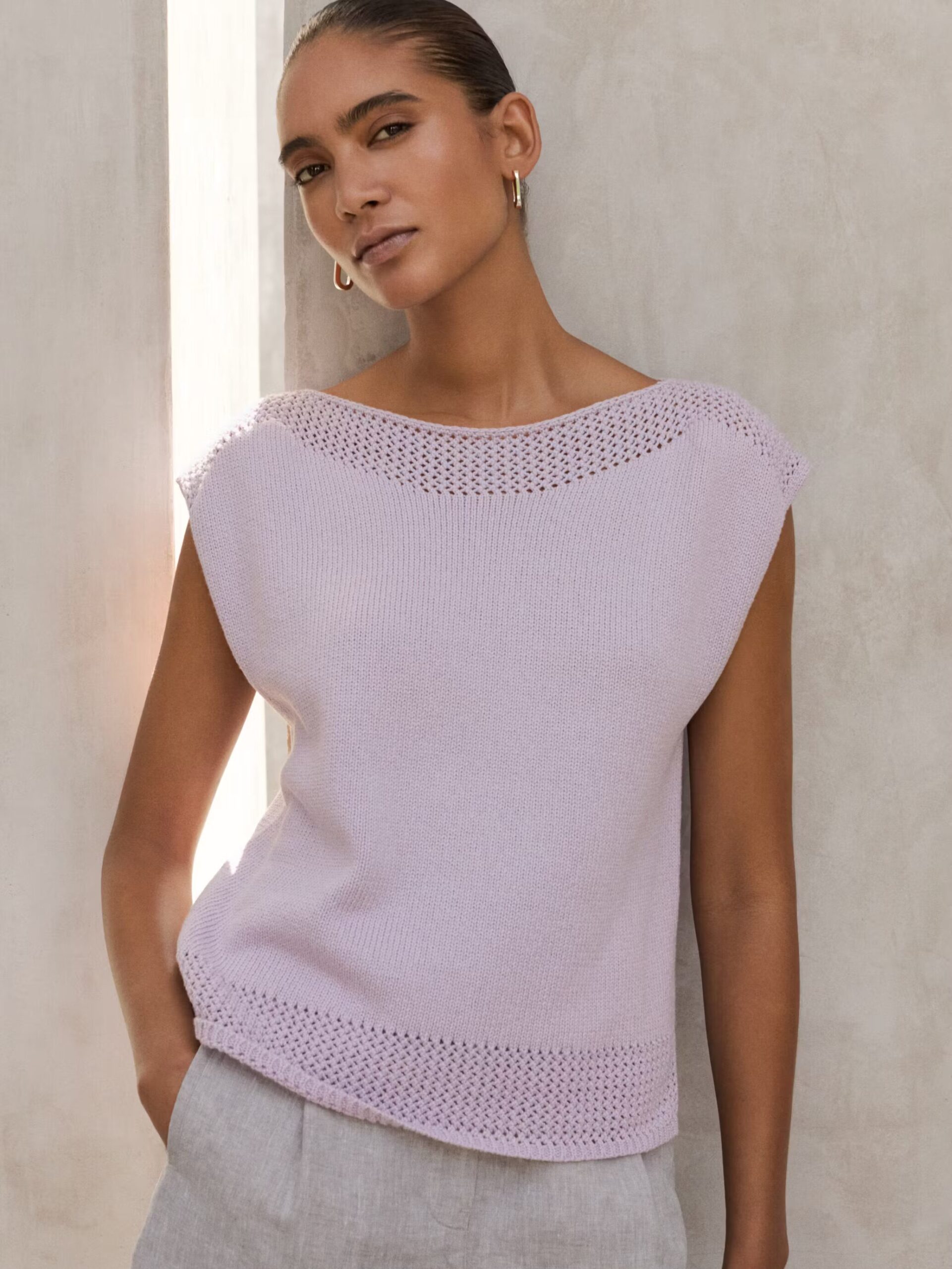 A woman wearing a sleeveless light purple knit top with eyelet details at the hem and neckline, paired with light gray pants, stands against a neutral textured wall.