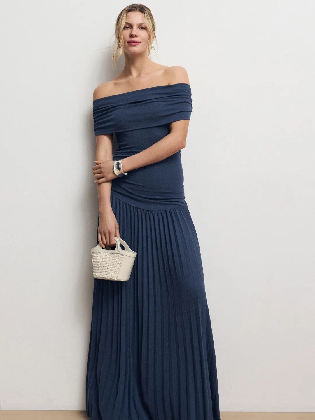 Woman standing against a plain wall, wearing an off-the-shoulder, floor-length blue dress, holding a small white handbag, and wearing sandals.