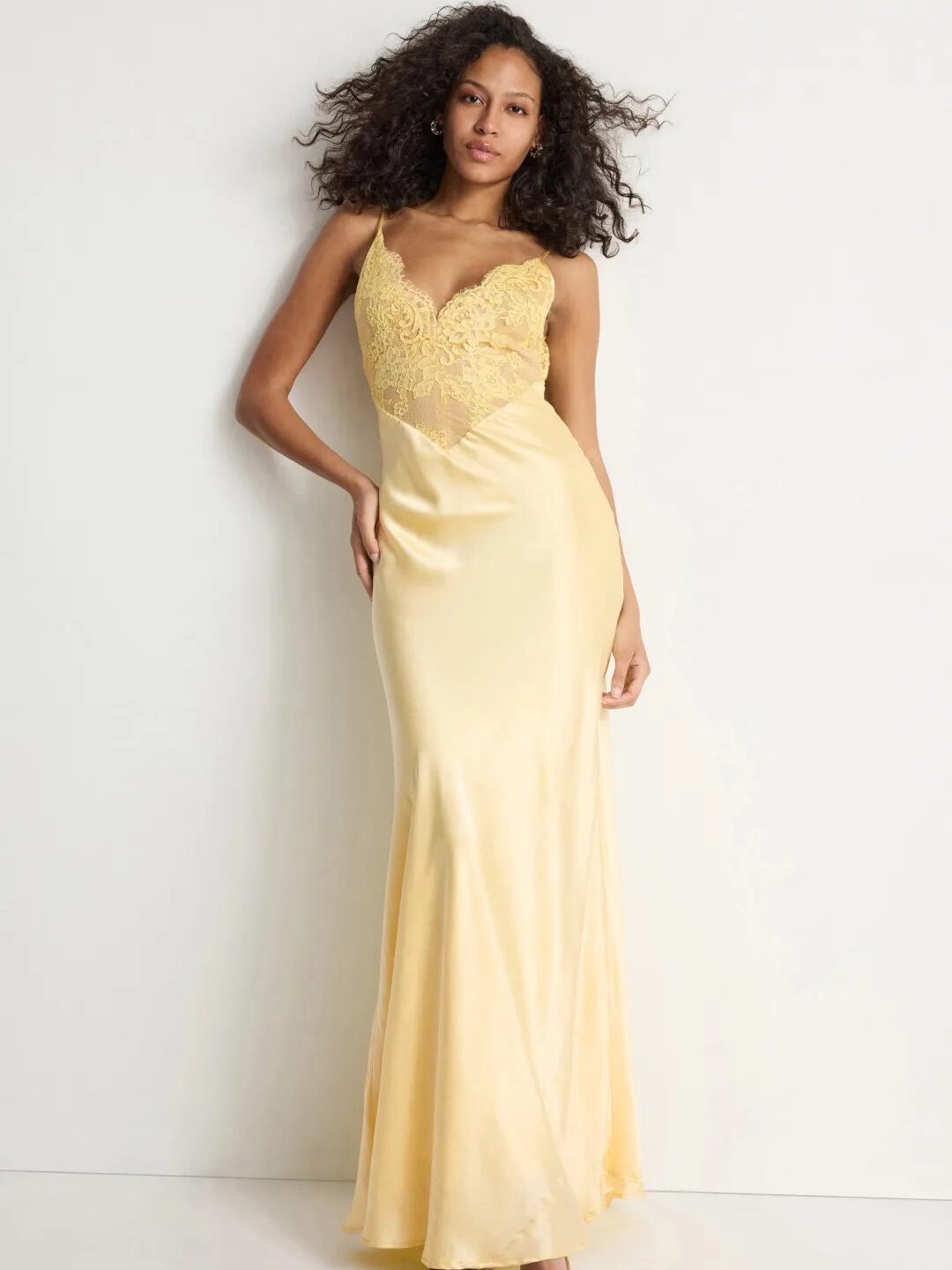 A woman stands against a white wall wearing a long, yellow satin slip dress with lace detailing on the bodice and open-toe heels.