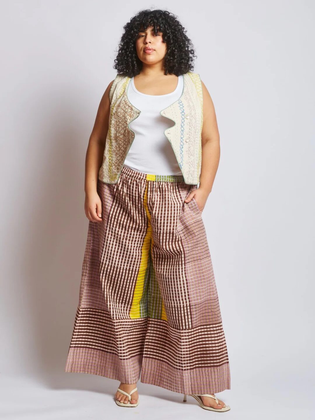 A woman stands against a plain background wearing a white tank top, a patterned vest, wide-legged checkered pants with yellow accents, and sandals.