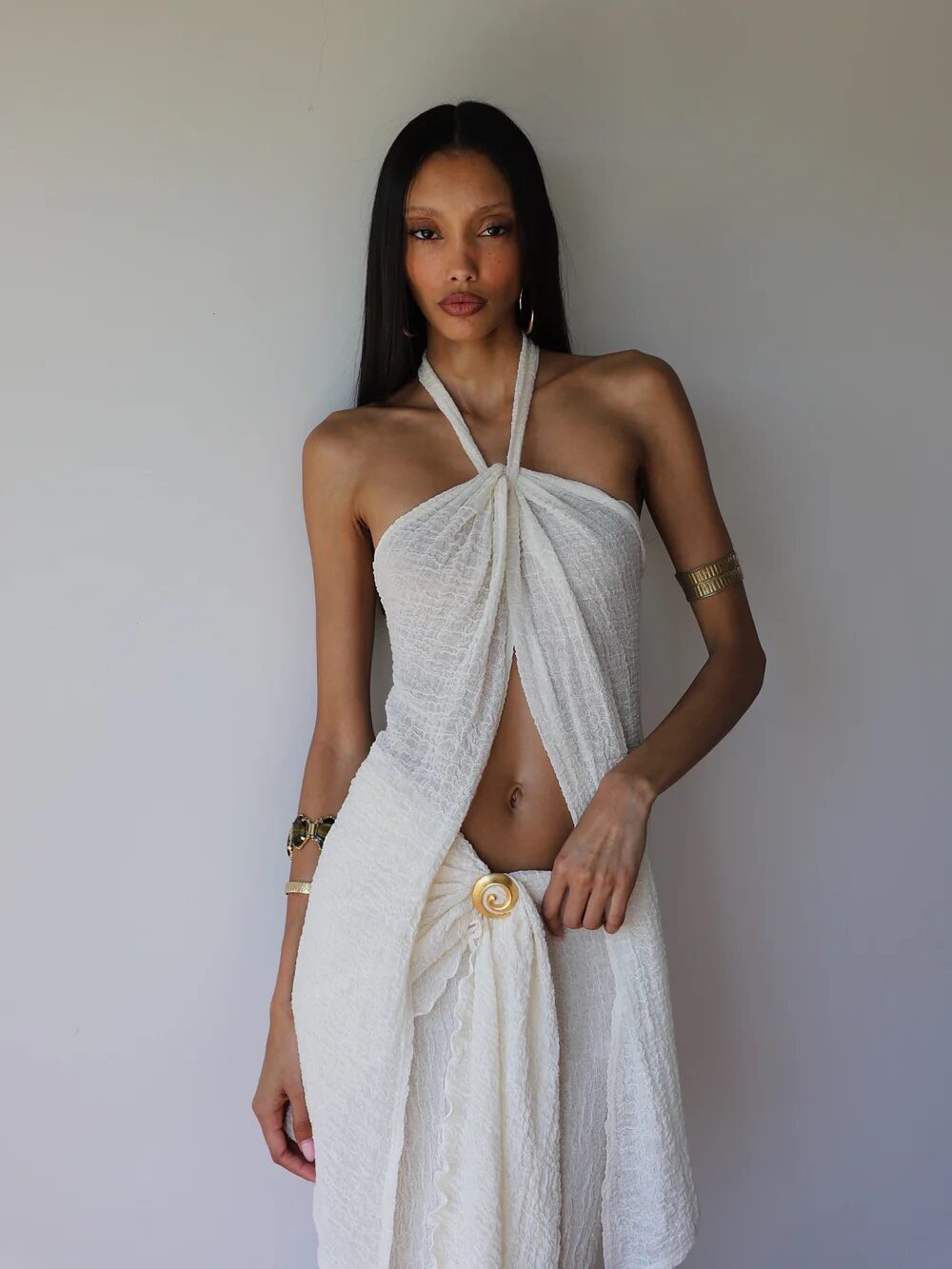 A woman wearing a white textured halter dress with a gold arm cuff and bracelet stands against a plain light gray background.