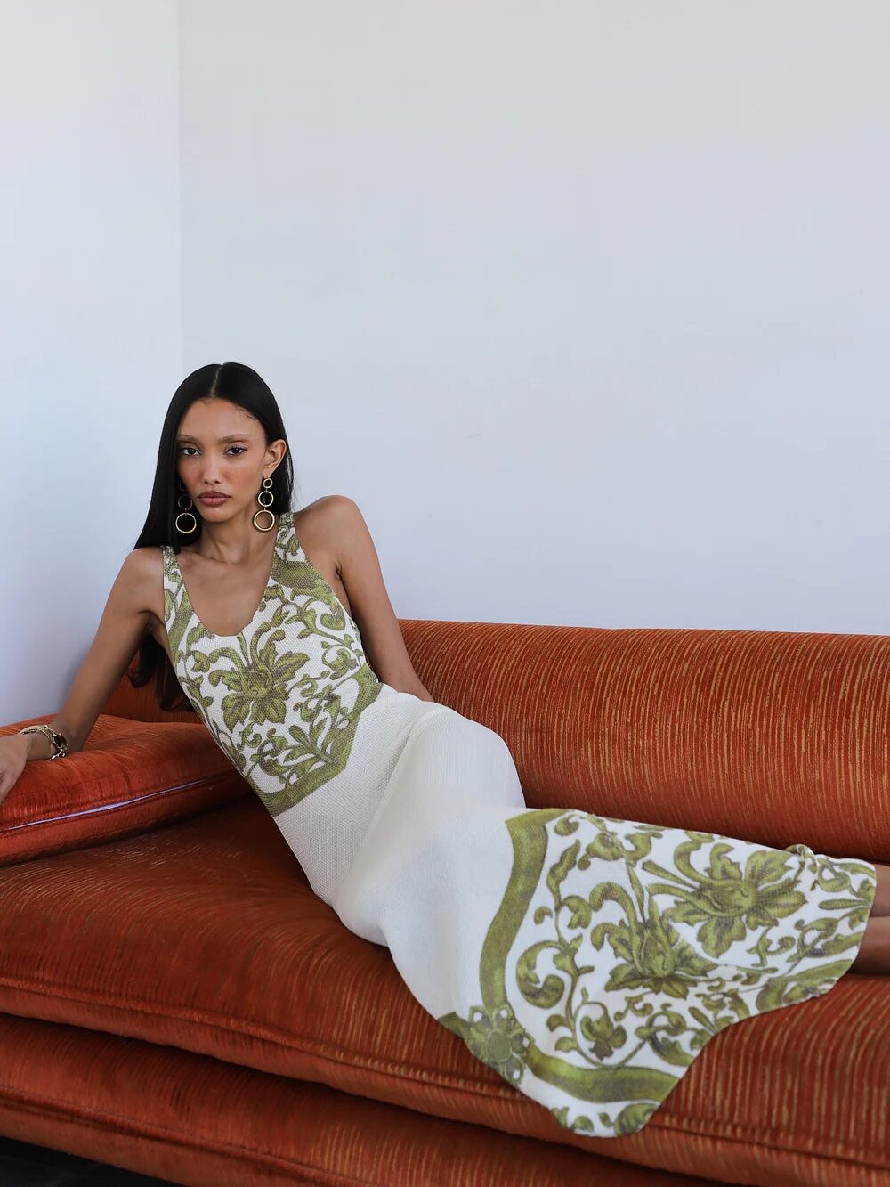 Woman in a long, sleeveless white dress with green floral patterns reclines on an orange sofa against a plain white wall.
