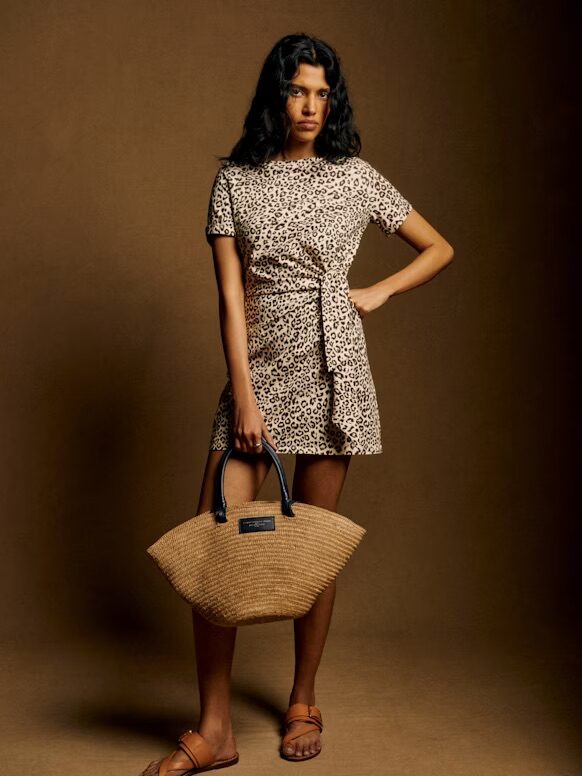 Woman stands against a brown backdrop wearing a short-sleeved, leopard print dress, brown sandals, and holding a woven beige handbag.