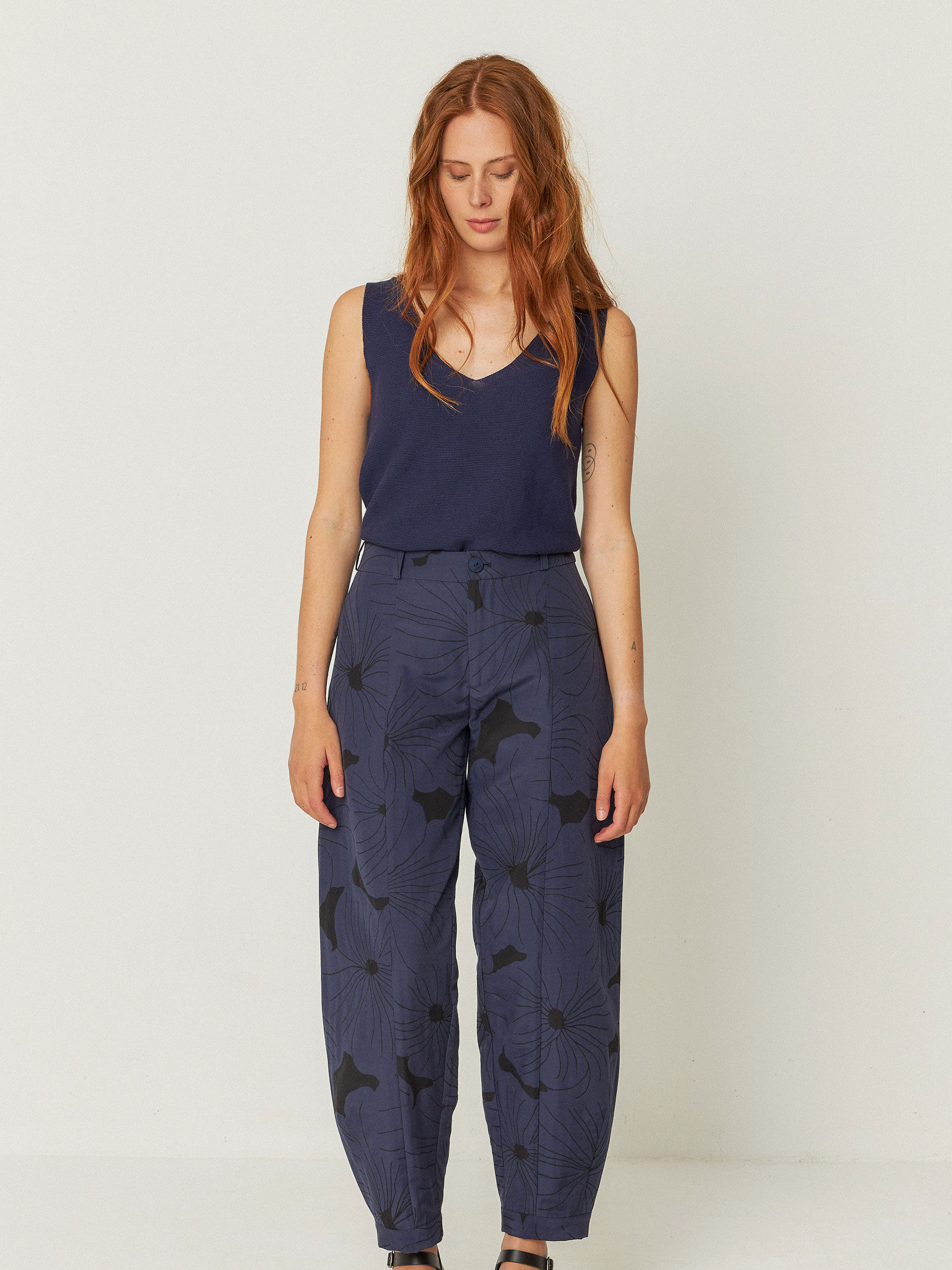 A woman with long red hair wears a sleeveless navy top, high-waisted navy floral print pants, and black sandals, standing against a plain light background.