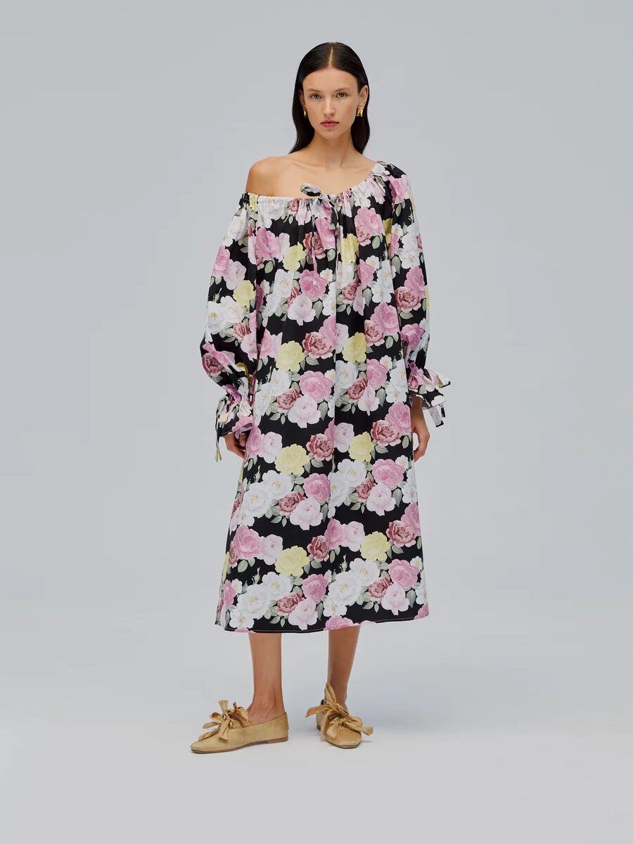 A woman stands facing forward wearing an off-the-shoulder, long-sleeve, floral midi dress with pink and yellow flowers, and tan shoes with bows against a plain light background.