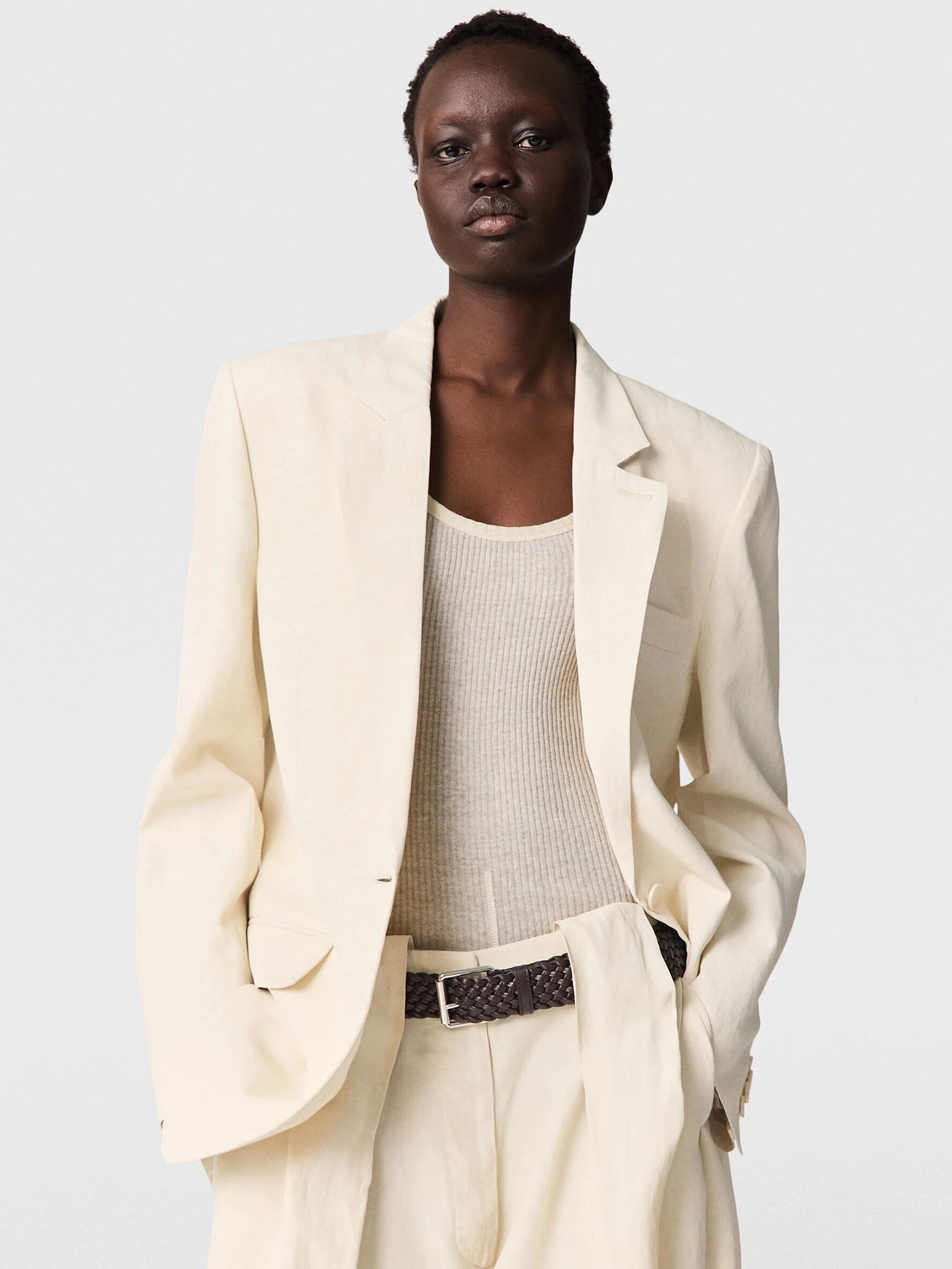 Person wearing a cream-colored suit with a matching tank top and a dark braided belt, posing against a plain light background.