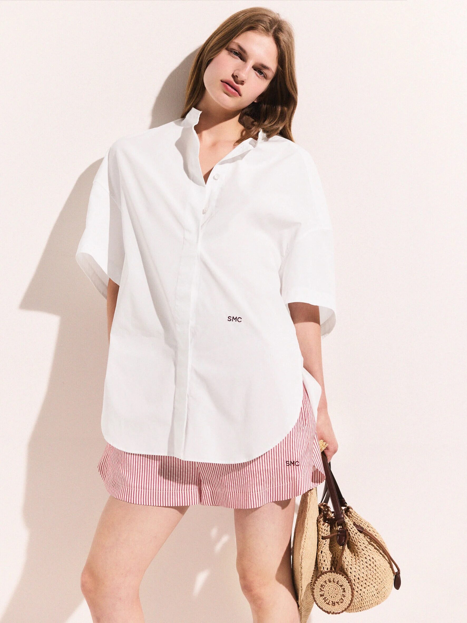 A woman stands against a light background wearing a white oversized shirt, red and white striped shorts, and holding a woven bag.