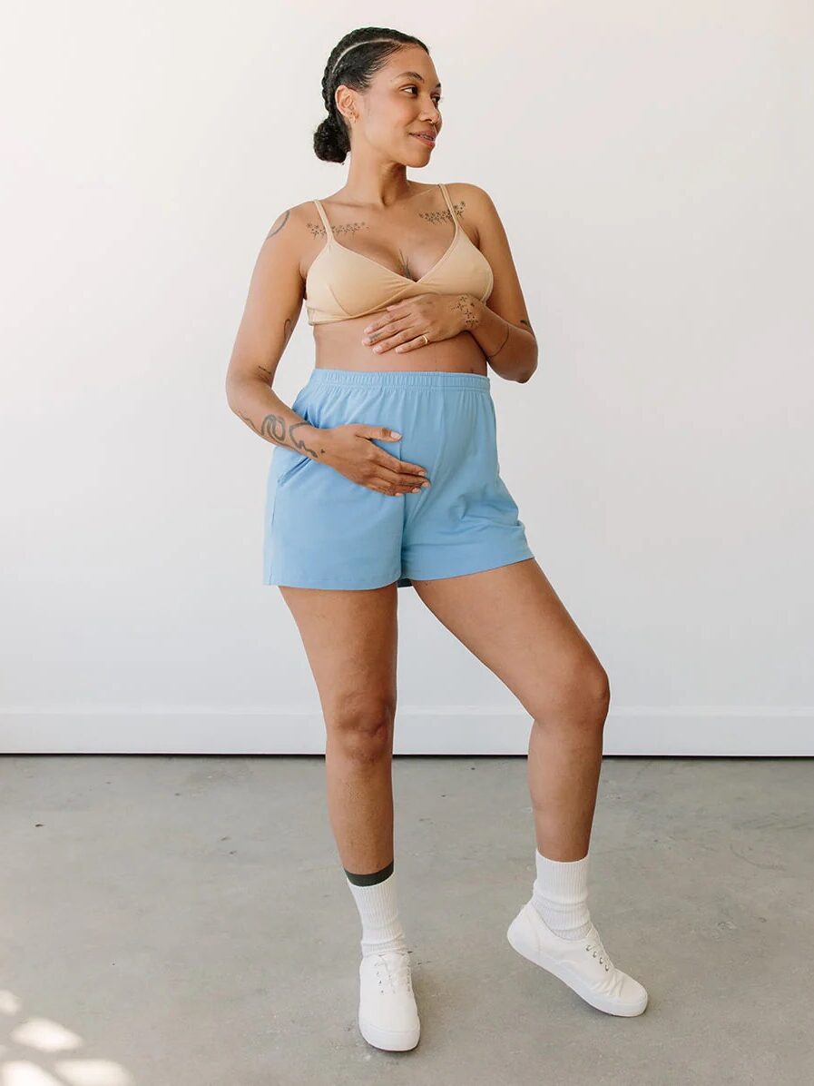 A pregnant woman stands indoors wearing a beige bra, blue shorts, white socks, and sneakers, with one hand on her belly and the other resting on her chest.