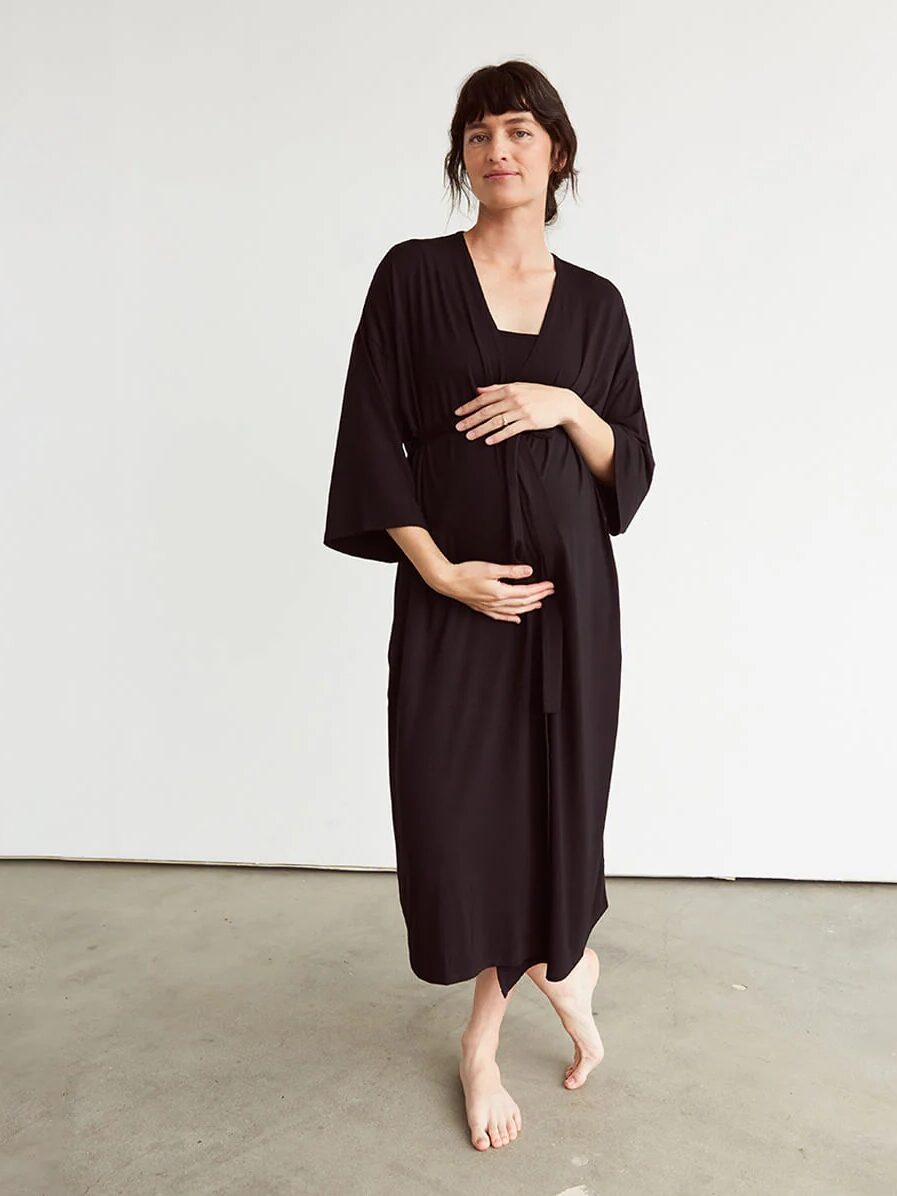 A barefoot pregnant woman stands indoors, wearing a black knee-length robe, with one hand resting on her belly and the other at her side.