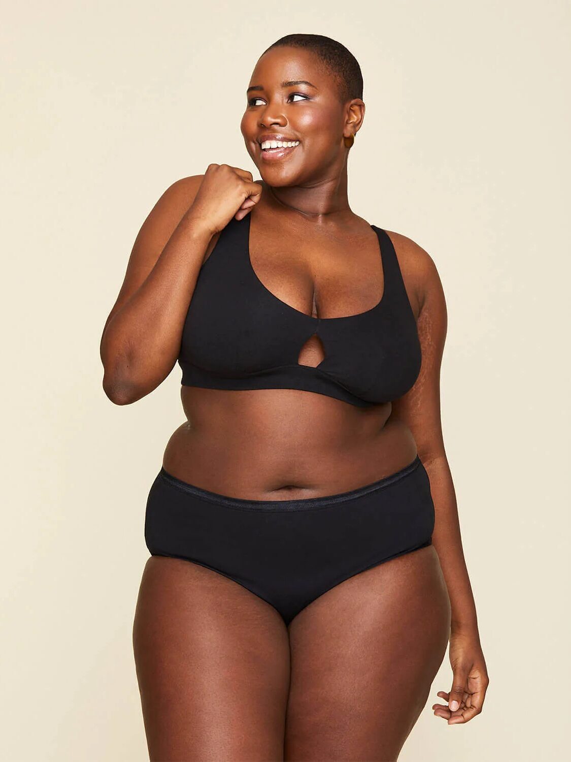 A woman stands smiling, wearing a matching black bra and underwear set, posing against a plain light background.