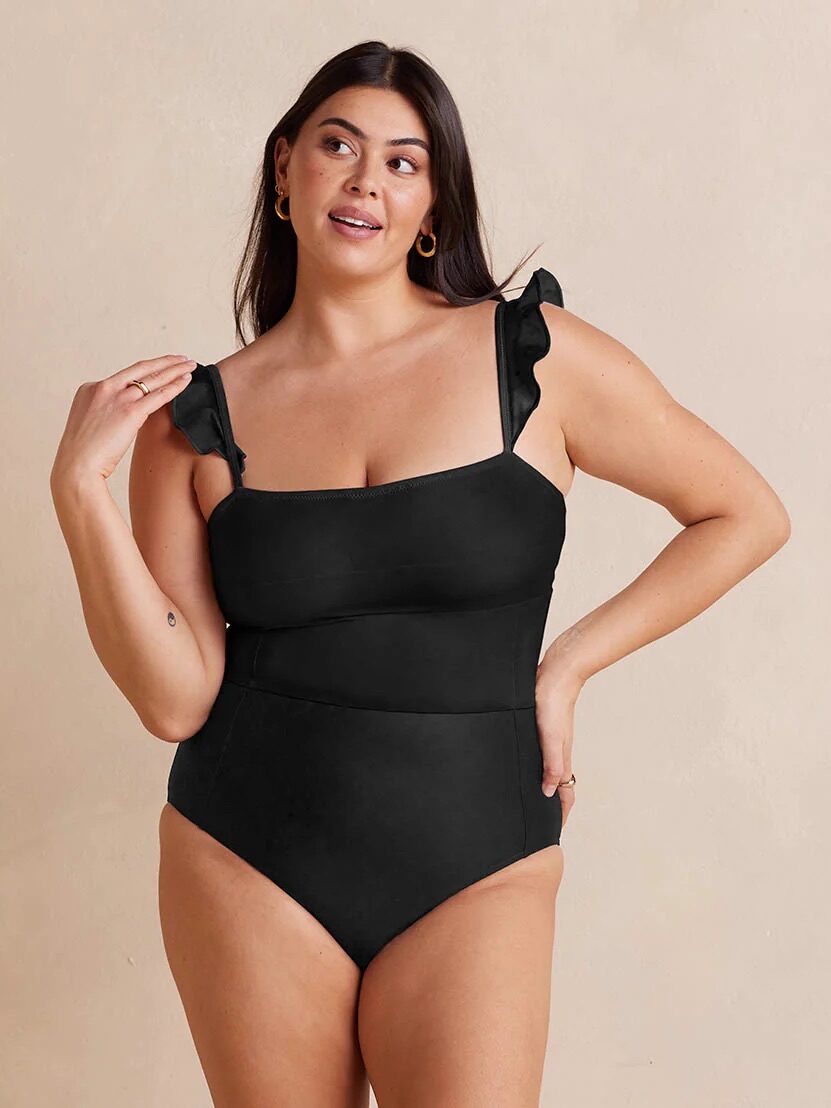 A woman poses in a black one-piece swimsuit with ruffled shoulder straps against a plain beige background.