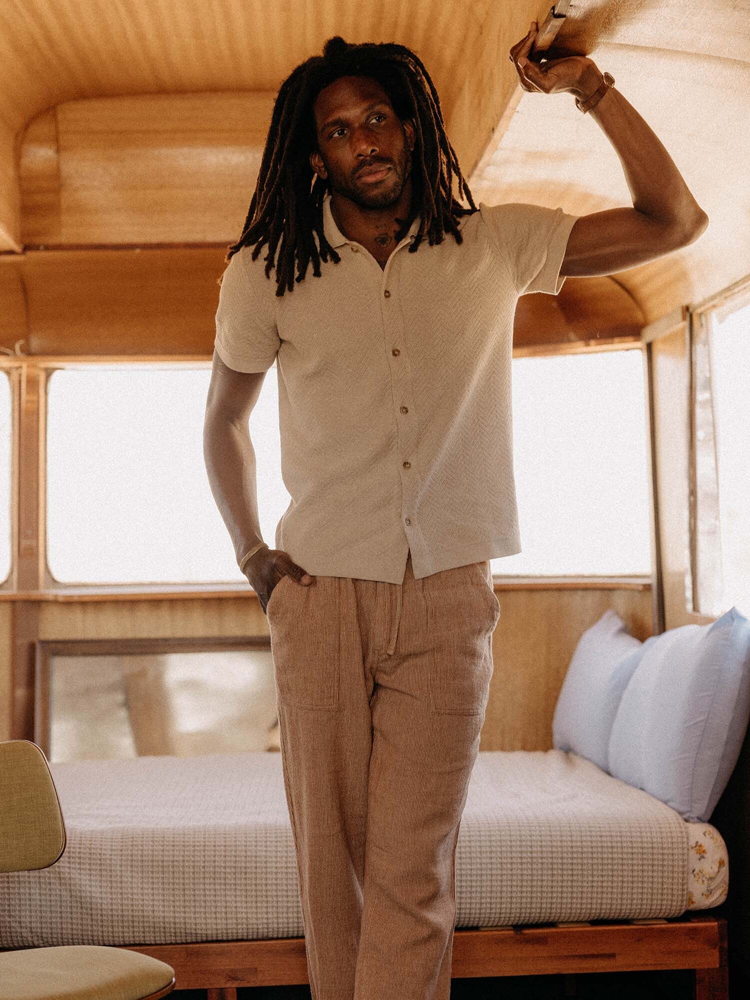 A man with long dreadlocks stands indoors, leaning with one arm against the ceiling near a bed in a warmly lit, wooden room.