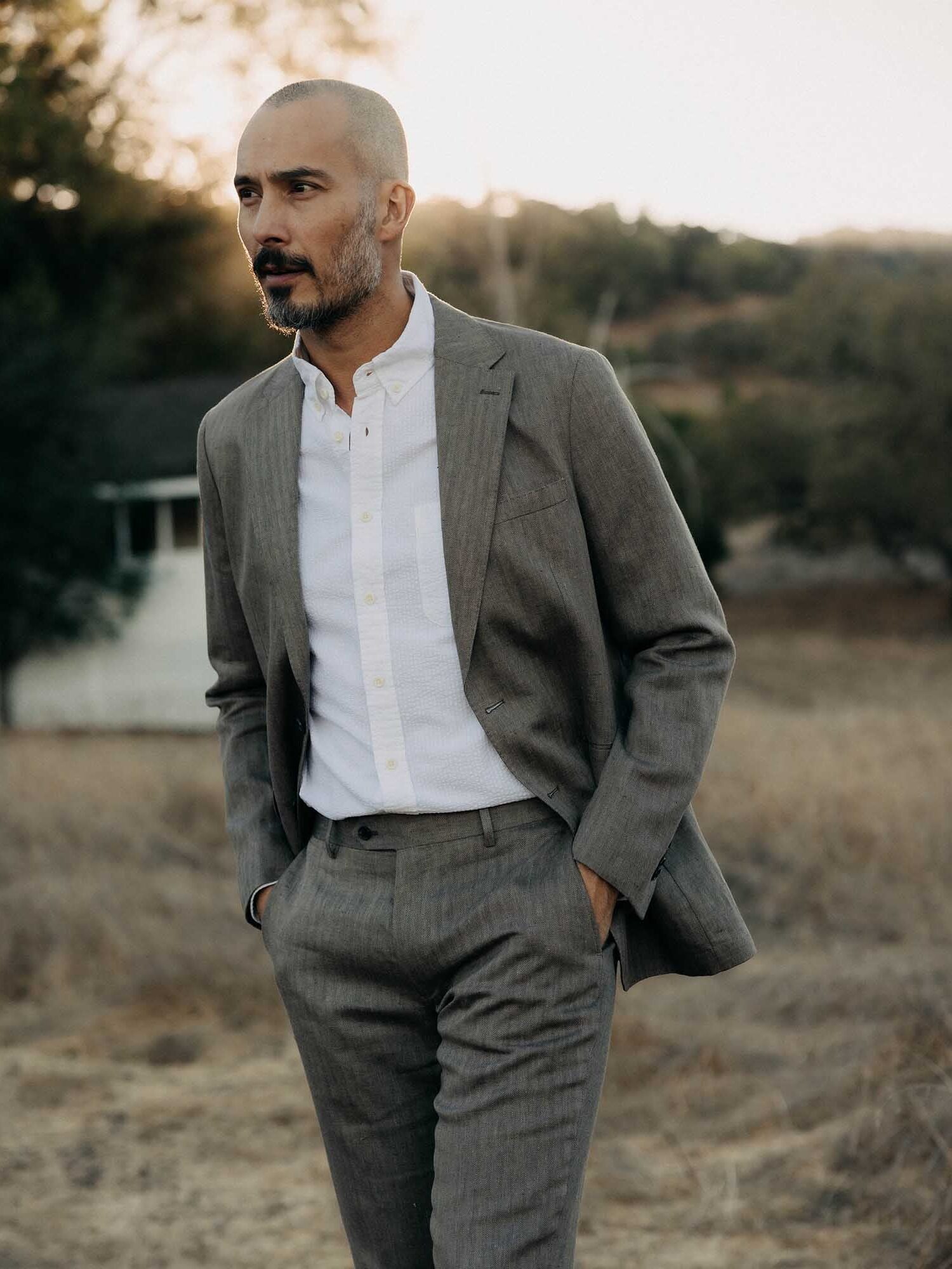 A man with a shaved head and beard wears a light gray suit and white shirt, standing outdoors in a dry, grassy landscape near a house.