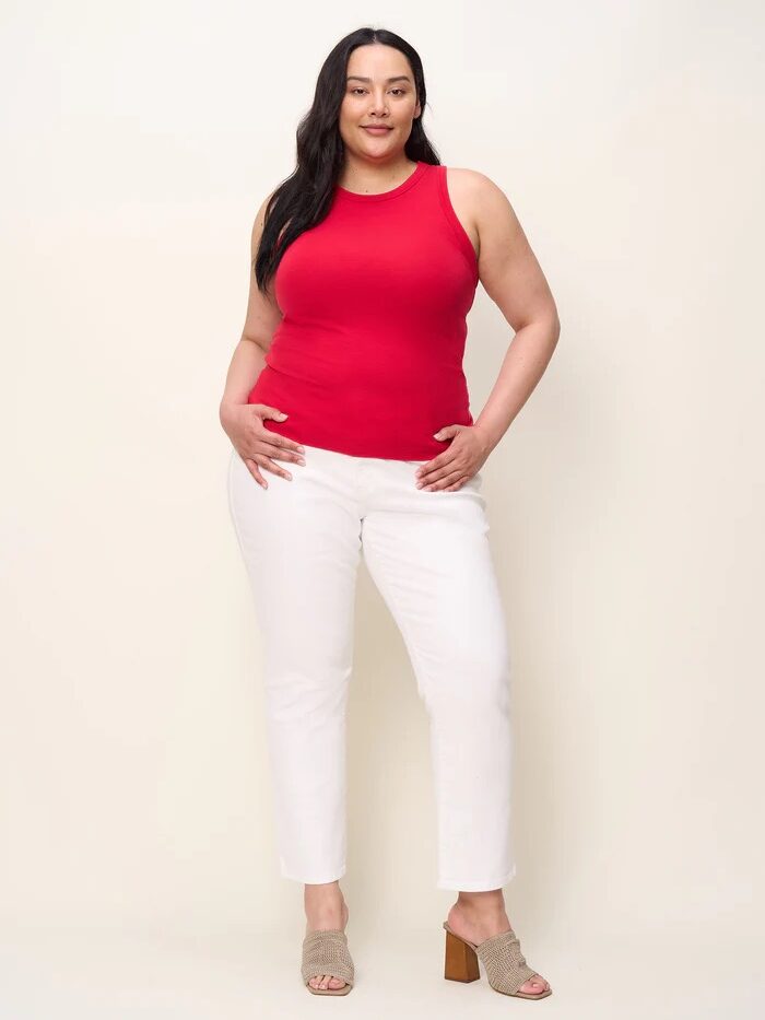 A woman stands facing forward, wearing a red sleeveless top, white pants, and beige heeled sandals against a plain light background.