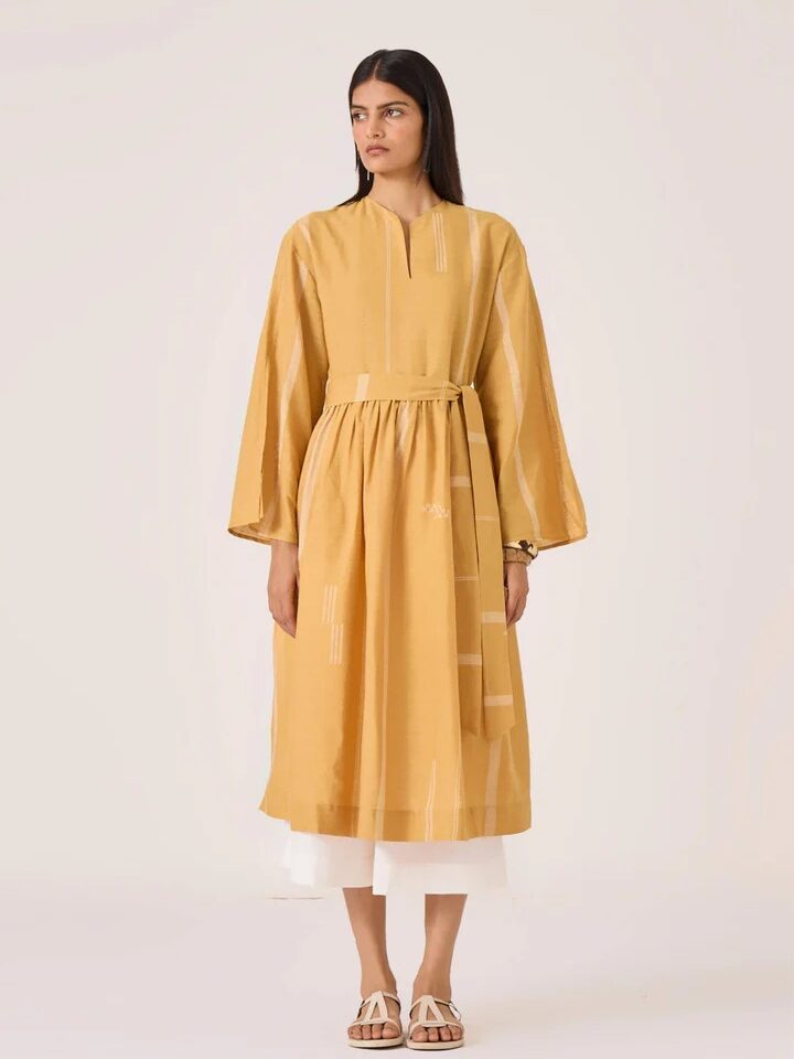 A woman stands wearing a mustard yellow, long-sleeved, belted dress with subtle patterns, layered over a white skirt and paired with white sandals.