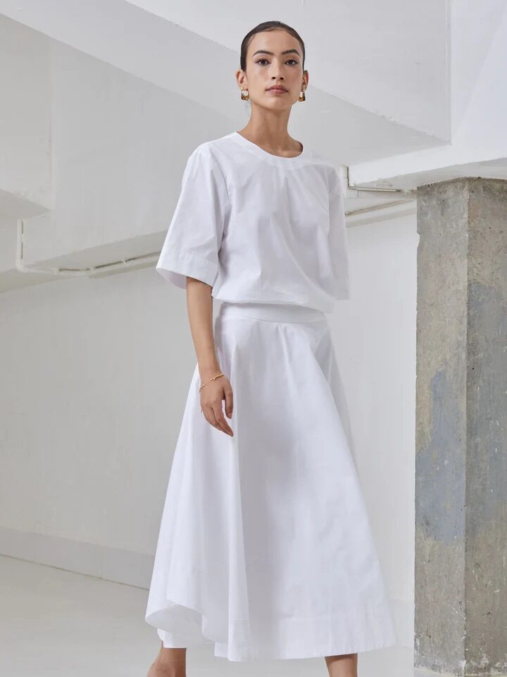 A woman wearing a loose white short-sleeve top, a white midi skirt, and beige heeled sandals stands indoors against a minimalist, white background with a concrete pillar.